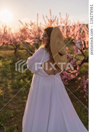 Woman blooming peach orchard. Against the backdrop of a picturesque peach orchard, a woman in a long white dress and hat enjoys a peaceful walk in the park, surrounded by the beauty of nature. Woman blooming peach orchard. Against the backdrop of a picturesque peach orchard, a woman in a long white dress and hat enjoys a peaceful walk in the park, surrounded by the beauty of nature. 116279561