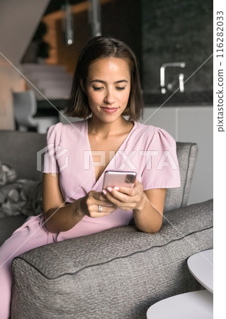 Positive young woman tying on mobile phone on home sofa 116282033