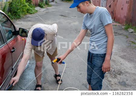 Dad and teenage son work together on the street near the garage. Outdoor activity. Family spend time. Helping and teach. Washing the car with a hand pump and hose. Bottled water. 116282358