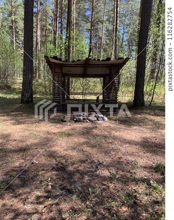Rustic wooden shelter in a pine forest with a prepared stone campfire pit inside the shelter. Rustic wooden shelter in a pine forest with a prepared stone campfire pit inside the shelter. 116282754