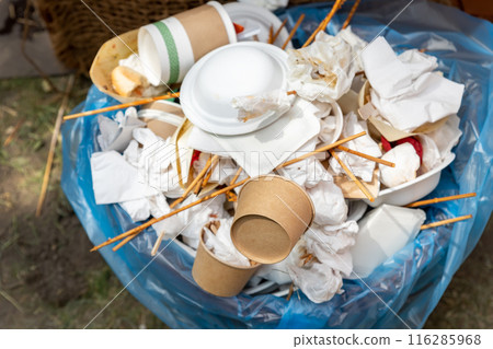 Close-up view of full garbage bin can with used dirty paper cardboard craft reusable sustainable cup, plates, towel napkins and cutlery at outdoor party park fest. Eco friendly recyclable tableware 116285968