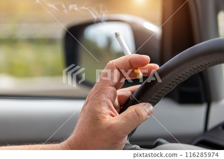 Close-up detail view of male hand driver holding cigarette while driving inside car on higway city street road on day time. Man person smoking inside vehicle. Low concentration attention danger Close-up detail view of male hand driver holding cigarette while driving inside car on higway city street road on day time. Man person smoking inside vehicle. Low concentration attention danger 116285974