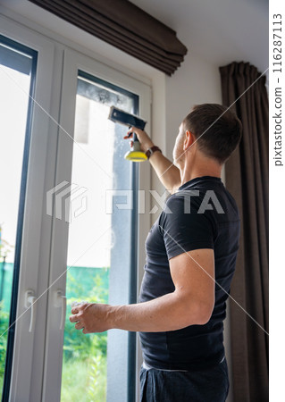 Young man cleans the windows in the house. He splashes detergent, rubs and removes water at the glass Young man cleans the windows in the house. He splashes detergent, rubs and removes water at the glass 116287113