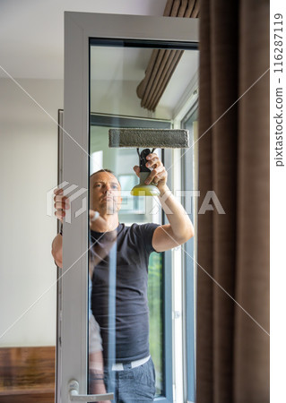 Young man cleans the windows in the house. He splashes detergent, rubs and removes water at the glass Young man cleans the windows in the house. He splashes detergent, rubs and removes water at the glass 116287119