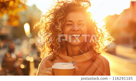 A beautiful woman with curly hair is smiling and holding a coffee in her hands against the background of the sun A beautiful woman with curly hair is smiling and holding a coffee in her hands against the background of the sun 116291185
