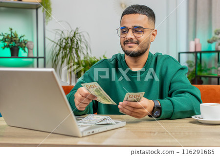 Smiling happy man counting money cash and use laptop pc calculate domestic income earnings at home Smiling happy man counting money cash and use laptop pc calculate domestic income earnings at home 116291685
