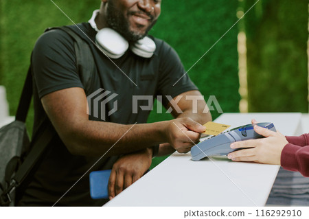 African American man tapping his golden credit card and smiling widely while female hand holding payment terminal African American man tapping his golden credit card and smiling widely while female hand holding payment terminal 116292910