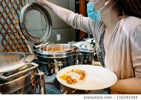 In a hotel setting, a woman is using a food warming machine to fill her plate at the buffet. In a hotel setting, a woman is using a food warming machine to fill her plate at the buffet. 116293304
