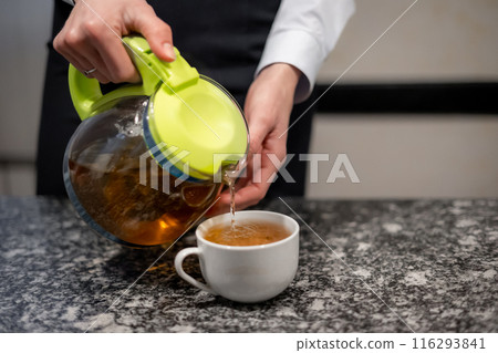 Close-up female hands pour tea from a glass teapot with a green lid into a white mug on marble table Close-up female hands pour tea from a glass teapot with a green lid into a white mug on marble table 116293841