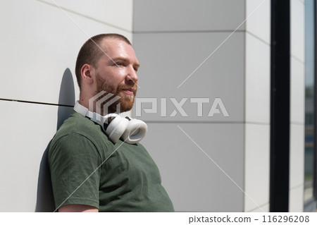 Portrait of caucasian handsome beard man wearing military color t-shirt with headphones near modern white wall listening music. Favourite song. Copy space. 116296208