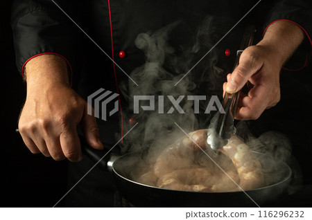 A cook turns a sausage with tongs in his hand while frying it in a frying pan. Low key concept of cooking sausage in the kitchen A cook turns a sausage with tongs in his hand while frying it in a frying pan. Low key concept of cooking sausage in the kitchen 116296232