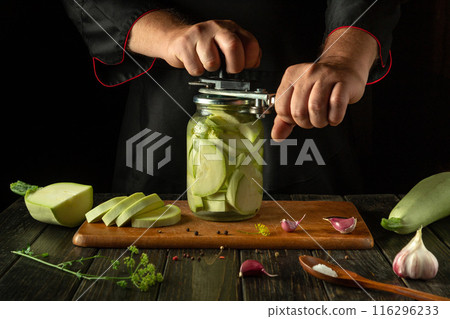 A cook uses a hand seaming tool to seal a jar of pickled zucchini on a kitchen counter. Vegetable marrow canning concept A cook uses a hand seaming tool to seal a jar of pickled zucchini on a kitchen counter. Vegetable marrow canning concept 116296233