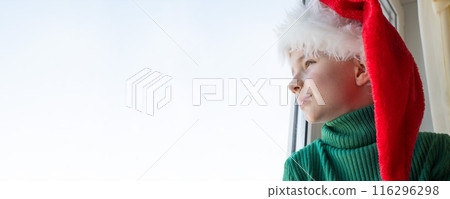 Children's dreams concept. Friendship. A lonely little boy in a Santa Claus hat is sad at the window waiting for gifts, looking out the window, the child is waiting for Santa Claus. The baby is 116296298
