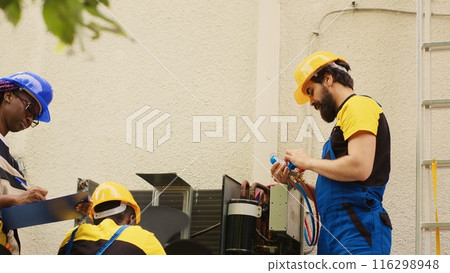 Adept repairmen diverse team working with manifold gauges to check air conditioner refrigerant tank in need of maintenance. Professional workers using barometer benchmarking hvac system component 116298948