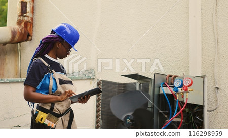 Qualified electrician servicing outdoor condenser internal components, checking freon level and calibrating thermostats. Precise serviceman imputing maintenance results data on laptop 116298959