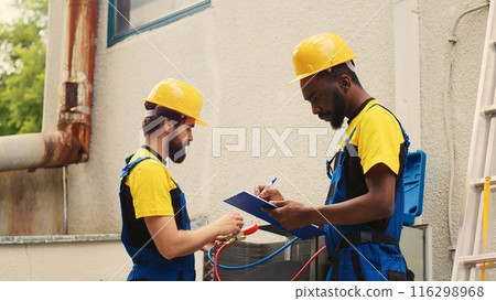 Seasoned mechanic doing condenser investigations, filter replacements and necessary fixes to prevent HVAC system failures while adept repairman coworker writes findings on clipboard 116298968