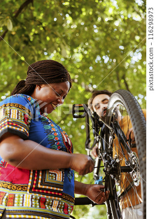 Sport-loving enthusiastic black woman fixing bike derailleur in yard for summer leisure cycling. Healthy and active african american female cyclist receiving help from caucasian man for bicycle 116298973
