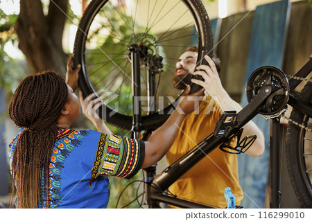 Energetic boyfriend and girlfriend dismantling damaged bicycle tire to repair and replace with new. Young enthusiastic multiracial couple doing maintanence on bike wheel in yard for leisure cycling. 116299010