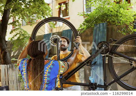 Young multiethnic couple happily attaching new bicycle tire for outdoor summer cycling. Black woman and caucasian man doing yearly inspection and maintenance on bike gear in yard. 116299014
