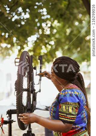 Young black woman repairing her own bicycle in yard, doing yearly maintenance of the vehicle with professional tools. Checking bike gear to fix, leisure activity during summer 116299016