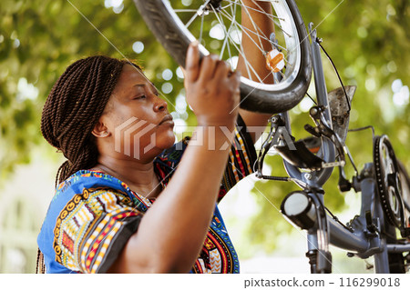 Energetic african american female dismantling damaged bicycle tire to repair and replace with new. Young athletic black woman grasping and carrying bike wheel for further maintenance. Energetic african american female dismantling damaged bicycle tire to repair and replace with new. Young athletic black woman grasping and carrying bike wheel for further maintenance. 116299018