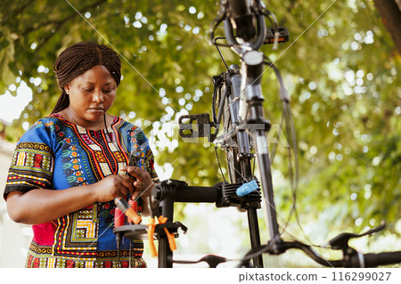 Young active black woman grasping a wrench for tightening screws and service bicycle parts outside. Sports-loving female holding professional tool for bike maintenance in home yard. Young active black woman grasping a wrench for tightening screws and service bicycle parts outside. Sports-loving female holding professional tool for bike maintenance in home yard. 116299027