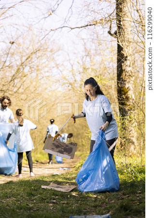 Woman environmentalist grabbing rubbish with a claw tool in the woods, clearing ecosystem of trash and plastic junk. Female activist doing voluntary work to tidy up the forest area. 116299040