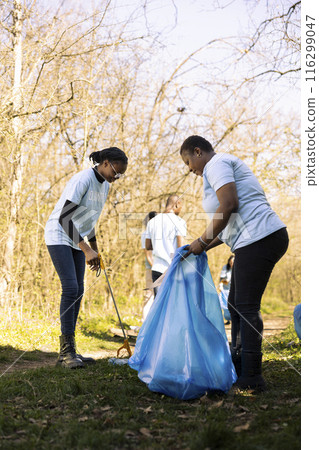 African american volunteers team working to tidy up the forest of rubbish, fighting pollution to protect the natural habitat. Women activists collecting trash and plastic junk with tools. 116299047