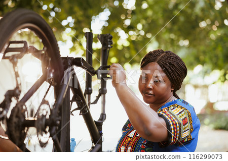 Young woman outdoors repairing bicycle tire with professional equipment. African american woman holding and checking bike wheel in yard for maintenance. Young woman outdoors repairing bicycle tire with professional equipment. African american woman holding and checking bike wheel in yard for maintenance. 116299073