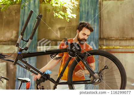 A fit young caucasian man repairs bike damage in home yard with professional repair-stand and tools. Dedicated athletic male engrossed in examining and fixing of modern bicycle outside with 116299077