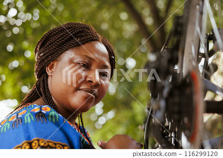 Closeup shot of athletic black woman meticulously examining and correcting damages on bicycle wheel chain. Image showing young female cyclist checking tire as annual bike maintenance routine. 116299120