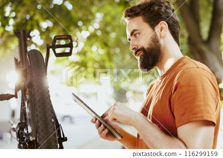 Image showing close-up view of young male cyclist holding smart device while servicing bicycle. Outdoorsy energetic caucasian man researching bike maintenance on the internet summer cycling enjoyment. Image showing close-up view of young male cyclist holding smart device while servicing bicycle. Outdoorsy energetic caucasian man researching bike maintenance on the internet summer cycling enjoyment. 116299159