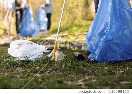Activist using claw tool to collect rubbish and plastic junk from the ground, storing garbage in a trash bag and clean the natural forest ecosystem. Male environmentalist gathering litter. Activist using claw tool to collect rubbish and plastic junk from the ground, storing garbage in a trash bag and clean the natural forest ecosystem. Male environmentalist gathering litter. 116299163