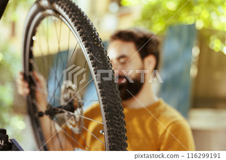 Close-up shot of bicycle wheel rubber carefully being examined and maintained by young healthy male in home yard. Image showing caucasian man adjusting bike tire for summer cycling. 116299191