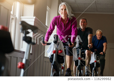 Woman, riding and bike at gym with earphone for fitness, training and workout listening to music for balance. Mature people, cycling machine and spin class exercise, wellness or cardio health goal 116299982