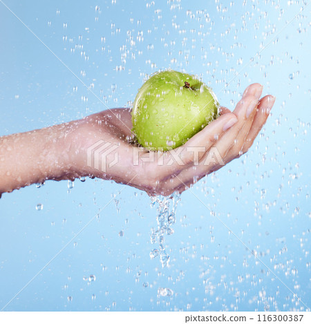Person, hand and water for apple in studio, washing and nutrition or sustainable skincare on blue background. Palm, cleaning and liquid for cosmetics or detox, antioxidants and snack or vegan fruit 116300387