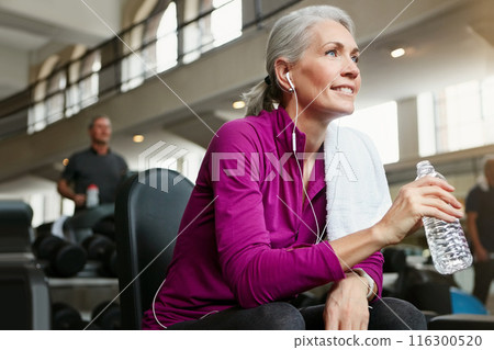 Mature woman, relax and drinking with water, earphones or thinking for music, workout or exercise at gym. Female person with smile for break, mineral beverage or thirst after training at health club 116300520