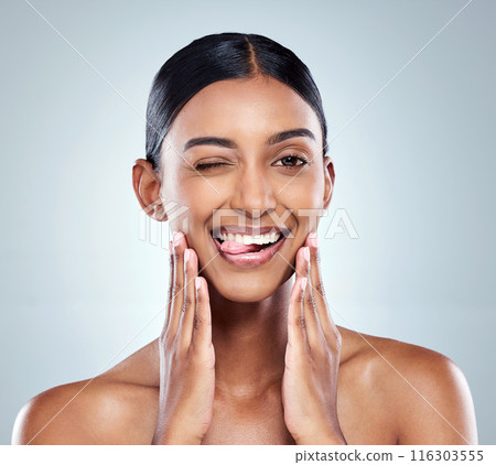 Portrait, wink and woman tongue with skincare for natural glow, skin health and shine results in studio. Indian girl, model and emoji face expression with beauty and dermatology by blue background Portrait, wink and woman tongue with skincare for natural glow, skin health and shine results in studio. Indian girl, model and emoji face expression with beauty and dermatology by blue background 116303555
