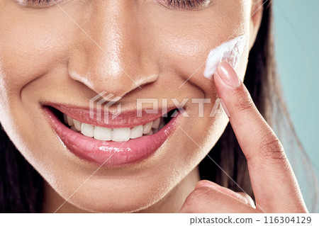 Woman, lotion and apply on cheek in studio, hydration and cleaning or skincare on blue background. Female person, beauty and cream for facial treatment or grooming, pamper routine and sunscreen 116304129