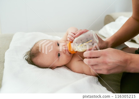 Hands of mother milk and feeding baby on sofa for growth development, nutrition or health. Drinking, woman parent and infant child with bottle for formula, wellness or bonding with love in home Hands of mother milk and feeding baby on sofa for growth development, nutrition or health. Drinking, woman parent and infant child with bottle for formula, wellness or bonding with love in home 116305215