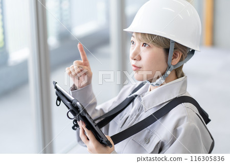 A young female worker with short blonde hair wearing a helmet and work clothes operating a tablet terminal A young female worker with short blonde hair wearing a helmet and work clothes operating a tablet terminal 116305836
