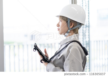 A young female worker with short blonde hair wearing a helmet and work clothes operating a tablet terminal A young female worker with short blonde hair wearing a helmet and work clothes operating a tablet terminal 116305837