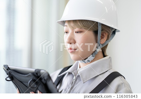 一名戴著頭盔、穿著工作服、留著金色短髮的年輕女工正在操作平板設備。 116305843