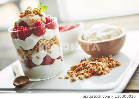 Yogurt, bowl and glass with fruit in cereal for breakfast food, nutrition and healthy meal on plate. Table, dairy and raspberry in container with wooden spoon for morning, organic snack or vegan diet Yogurt, bowl and glass with fruit in cereal for breakfast food, nutrition and healthy meal on plate. Table, dairy and raspberry in container with wooden spoon for morning, organic snack or vegan diet 116306036