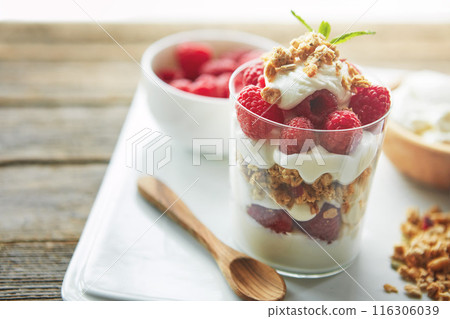 Glass, cereal and yogurt with raspberry with spoon for vegan food, organic or meal at breakfast. Fiber, dairy and fruit on plate by clear container with protein for morning, healthy nutrition or diet 116306039