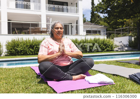 Outdoors, senior biracial woman sitting cross-legged on a yoga mat, meditating 116306300