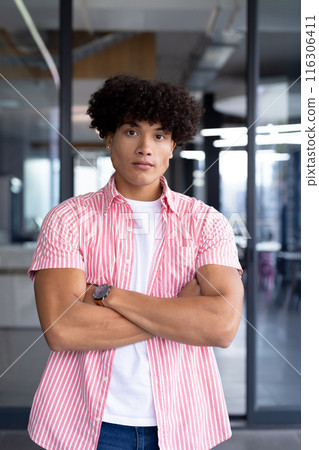 In modern office, a young biracial male coworker standing with arms crossed 116306411