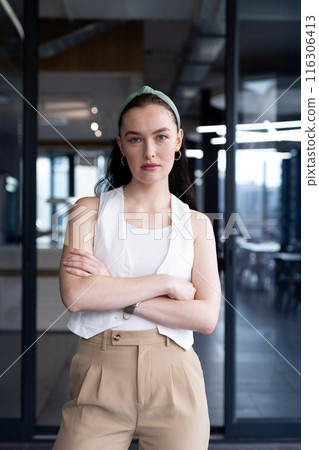 Young Caucasian female in casual office attire stands, arms crossed in modern office 116306413