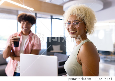 In modern office, two biracial coworkers holding coffee, laughing together 116306431
