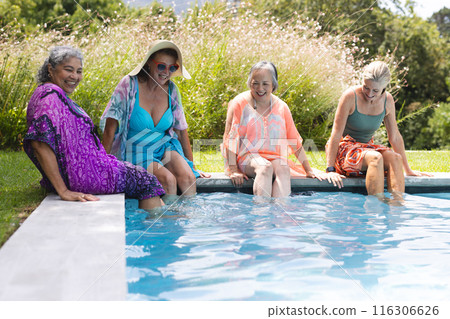 Outdoors, diverse senior female friends sitting by pool, dipping feet in water, copy space Outdoors, diverse senior female friends sitting by pool, dipping feet in water, copy space 116306626